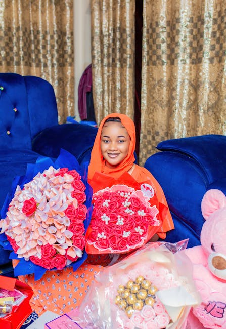 A smiling woman wearing an orange hijab and matching top holds two heart-shaped floral arrangements made of light pink and deep pink roses, with additional flowers and decorative elements, in a cozy i