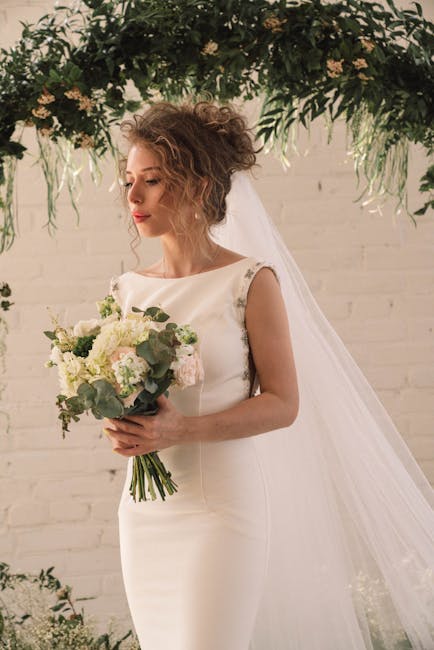 A bride with curly, voluminous hair styled in an elegant updo, wearing a sleeveless, ivory wedding gown with a high neckline, standing indoors against a white brick wall adorned with lush greenery and