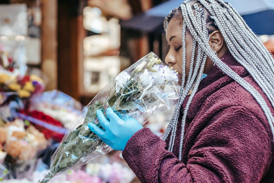 A woman with silver braids wearing a burgundy fleece jacket and blue gloves is at a flower market, inspects a bouquet of white roses wrapped in clear plastic, with various other flowers and floral arr