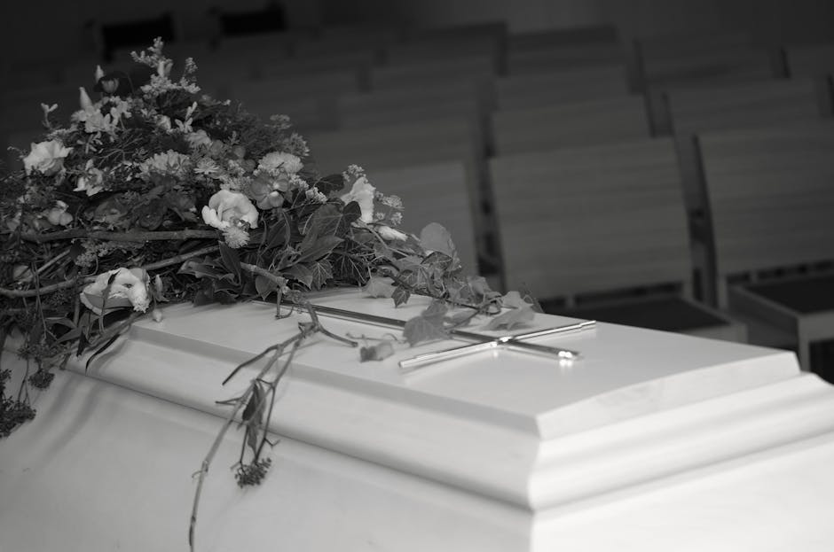 A white coffin adorned with an arrangement of fresh flowers, including roses and greenery, resting on top at a funeral service. The flowers are wrapped loosely with some petals and stems extending ont