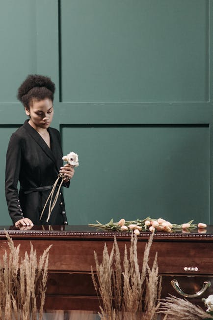 A mournful woman with dark curly hair styled in a neat updo, dressed in a black blazer, holds a small white flower bouquet in her left hand while standing beside a polished dark wooden casket. On the 