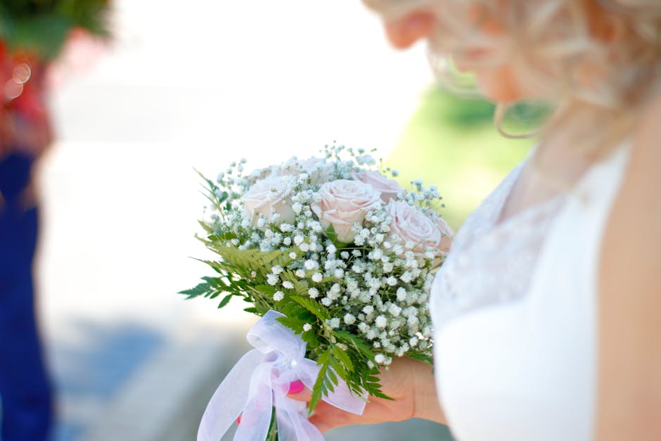 A close-up of a wedding bouquet featuring soft pink roses, delicate white baby's breath, and lush green foliage, wrapped with a white ribbon. The bouquet is held by a bride dressed in white, with a bl