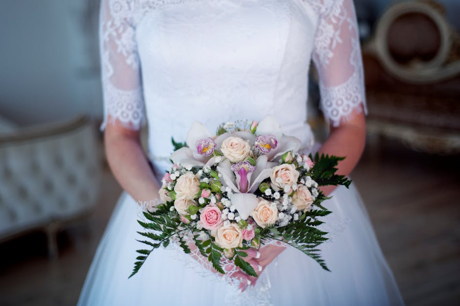 A bride holding a bouquet arrangement featuring pale pink roses, white calla lilies, and delicate pink and yellow orchids, accented with small white baby's breath and lush green ferns. The bouquet is