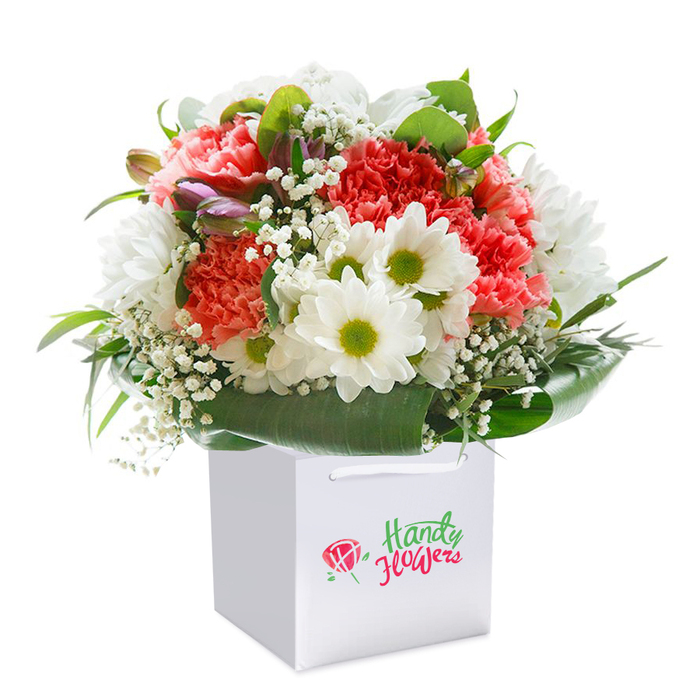 A vibrant, expertly crafted bouquet sits proudly within a structured square white gift bag, its crisp silhouette highlighting the artistry and freshness beloved by flower lovers throughout Merton. At its heart, generous coral-pink and red carnations unfurl in profuse, ruffled layers, creating a tapestry of warm colour and touchable softness, much like the displays seen at local flower markets near Merton Abbey Mills. Spotless white daisy chrysanthemums, their vivid lime-green eyes alight, add sparkle and charm, while clusters of delicate baby's breath float gently throughout, the epitome of weightless elegance. The bouquet is framed by an ensemble of lush, broad green leaves sweeping out and encircling the base, giving the whole composition a beautiful symmetry and balanced fullness. Amidst the main blooms, a few closed buds-hinted with purples and pinks-emerge quietly, promising renewed life and a thoughtful eye for detail. The sturdy white handles of the gift bag are built for easy transport-whether you're delivering this uplifting arrangement across the hills of Cannon Hill Common or presenting it at a family celebration in Lower Morden. Soft, even light flatters every petal and leaf, underscoring the bouquet's sense of freshness and skilled curation. This arrangement is an ideal choice for anyone seeking a memorable and heartfelt floral gift in Merton-perfect for birthdays, thank-yous, or simply sending joy to a cherished friend or neighbour.