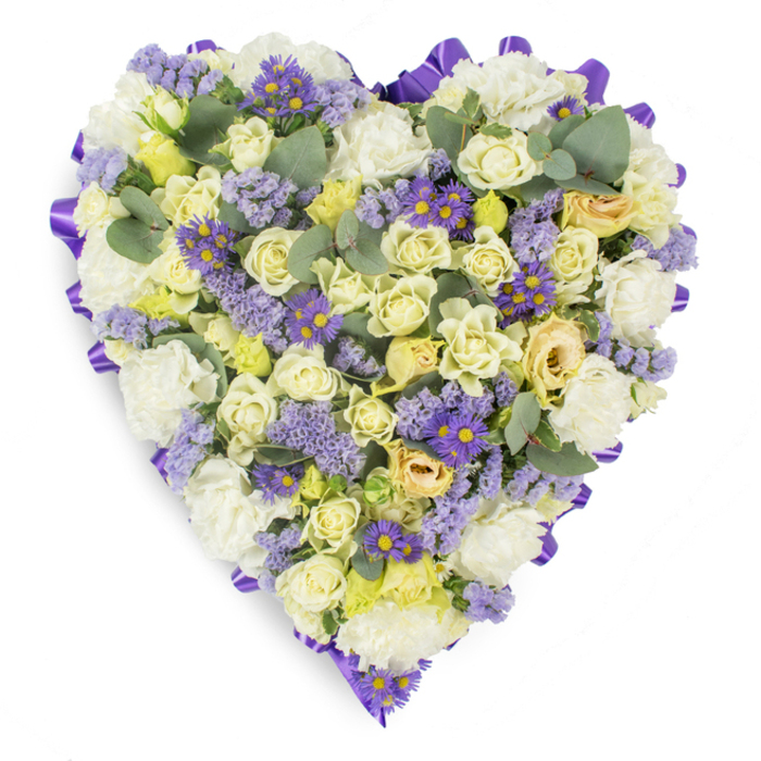 A meticulously arranged heart-shaped floral tribute set on a stark white backdrop, this design balances softness with clear structure. The heart's interior is dominated by tightly clustered cream and pale yellow roses-some in bud, others softly unfurling-giving the piece a layered, tactile depth that invites quiet contemplation. Interspersed among the roses are generous clusters of white carnations, their ruffled edges adding volume and a time-honoured, comforting presence. Lighter tones are contrasted by sprigs of delicate lavender statice that float lightly across the surface, and by small, vivid purple aster-like blooms with bright yellow centers that create eye-catching points of color and texture. Scattered throughout are rounded eucalyptus leaves in a cool blue-green, their smooth, matte surfaces breaking the floral mass and suggesting a subtle, refreshing aroma. The whole heart is bordered by a neat, continuous purple satin ribbon, folded into a looped edge that defines the silhouette and lends a refined finish appropriate for formal memorials. Photographed in soft, even light to emphasize the subtle tonal shifts, the composition conveys peace, respect, and lasting affection. Suited for funeral services, wakes, or graveside placement in Merton and surrounding areas such as Mitcham and Raynes Park, this heart-shaped arrangement communicates sympathy and devotion with understated elegance.