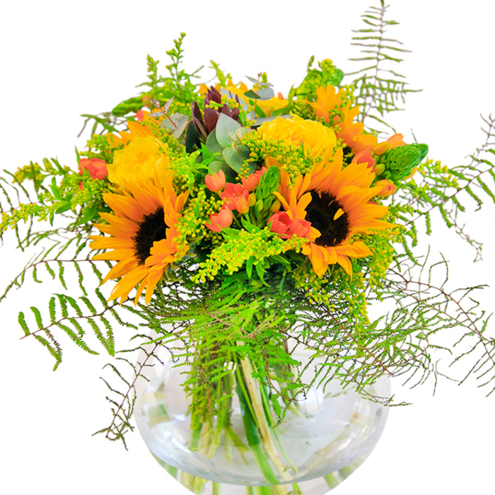This spirited bouquet mirrors the energy and warmth that courses through Merton at the height of the season. Centered on dramatic sunflowers, their wide, vibrant yellow petals flaring around deep brown cores, the arrangement commands attention much like the lively festivals along Morden Road. Richly-textured yellow garden roses unfurl their inner whorls, the cream-to-gold gradients echoing the charm of local English gardens. Between these, clusters of shiny red hypericum berries are dotted throughout, injecting playful flashes of red that evoke the dynamic spirit of Merton's friendly community gatherings. Draped greenery cascades in feathery layers-fern fronds and goldenrod lend subtle movement, texture, and a countryside freshness, while the blue-green eucalyptus threads understated coolness through the composition. The rounded glass vase, water shimmering inside, provides a modern and clean setting, allowing each stem to display its unique character while complementing the bouquet's lively, summery palette. Light glances off the petals, creating a cheerful interplay of shadows and brilliance that mimics a bright day in Cannon Hill Common. Ideal as a birthday or anniversary gift, for same-day delivery around Merton, or as an uplifting centrepiece for local family dinners, this bouquet reflects both the artistry of its creator and the inviting personality of the area it calls home.