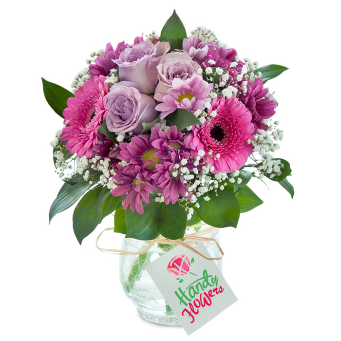 Taking inspiration from the friendly, vibrant character of Merton, this meticulously assembled mixed bouquet glows within a subtly crackled, clear glass vase against a pure white backdrop. The arrangement is an inviting harmony of pink, purple, and lavender, centered on a cluster of pale lavender roses whose petals seem to spill open with refined beauty and a gentle, sweet scent. Large, confident magenta Gerbera daisies stand out, their open faces and dark centers capturing the eye with joyful exuberance, reminiscent of celebrations at Wimbledon Village. Interwoven between them, diminutive yet vivid plum chrysanthemums with greenish-yellow hearts add texture and depth, creating a playful interplay with sprays of snow-white baby's breath. Each delicate gypsophila floret adds a touch of soft airiness, brightening the entire composition and evoking memories of tranquil days in Merton Park. Glossy green leaves anchor the bouquet, emerging with a freshness that feels garden-picked and spontaneous. The vase's textured finish sparkles in the light, while a single strand of rustic raffia ties round the neck, holding a blank cardstock tag ready for your personal note-an understated invitation to mark birthdays, express thanks, or simply share a joyful surprise. This arrangement, typical of Merton's expert floral style, is ideal for thoughtful local delivery-bringing lively colour, elegant craftsmanship, and a dose of happiness to any doorstep in Raynes Park or South Wimbledon.