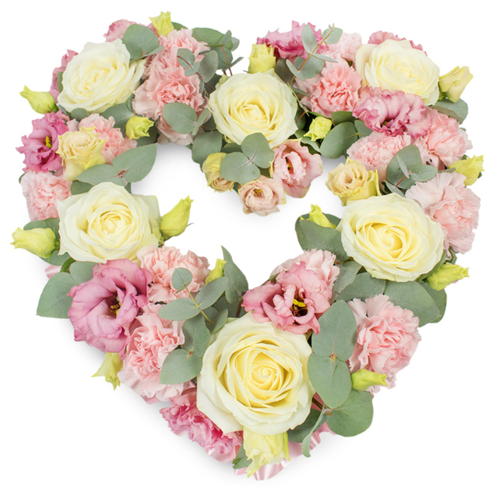 A full, heart-shaped floral tribute fills the frame against a crisp white background, its pastel flowers carefully arranged in a design that would sit gracefully in a Merton church, crematorium chapel, or family home during a time of farewell. The heart outline is perfectly formed and completely covered with blooms, giving a sense of completeness and deep devotion. At its core are several large cream and pale yellow roses, their velvety petals spiralling outward from tightly furled centres into generous rounded blossoms. These roses act as anchors, creating calm focal points that draw the eye around the entire heart. Interspersed among them are clusters of light pink carnations, each head filled with frilled, delicately overlapping petals that provide a soft, cushion-like texture. Scattered throughout the design, deeper pink to mauve lisianthus blooms open into graceful cups, their slightly ruffled edges adding gentle movement and a more intense note of colour, like the last light over Wimbledon Park at the end of the day. Silvery-green eucalyptus leaves wind naturally through the arrangement, their round and oval shapes peeking through the petals and outlining the heart with a cool, soothing green tone. Here and there, small yellow-green buds emerge, symbolising fresh growth and hope. The flowers are packed densely, giving the wreath a plush, luxurious feel, as though every stem has been placed with thought and affection. This heart-shaped arrangement evokes love, remembrance, and sympathy, making it a fitting choice for funerals, memorial services, or commemorative anniversaries across Merton, where families seek something gentle yet expressive to honour a loved one's memory.
