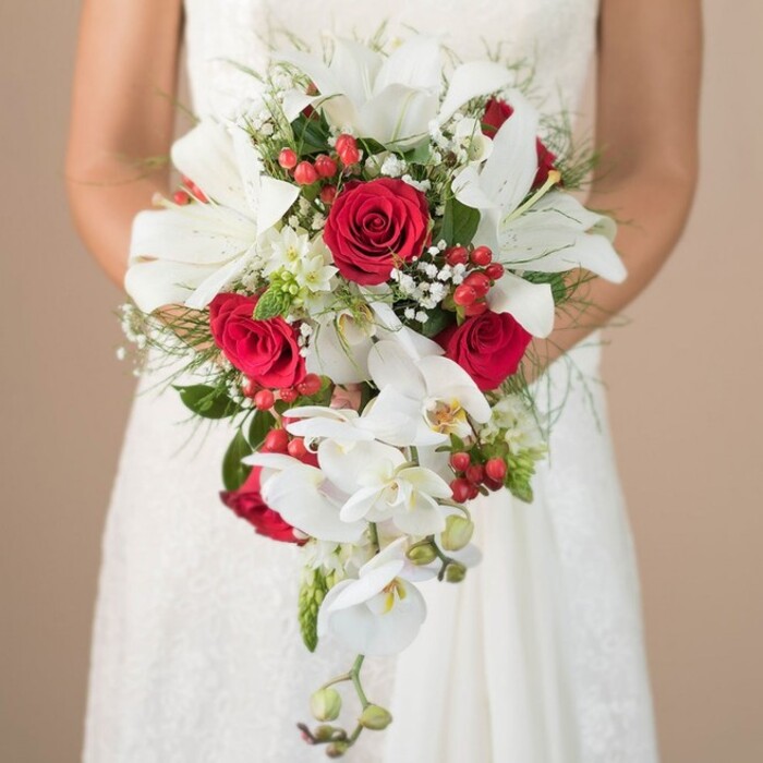 Set against a softly blurred, warm-toned background, a bride from Merton is captured waist-up, the focus drawn to the exquisite teardrop bouquet she holds. Radiant white oriental lilies, each broad petal basking in gentle light, sit prominently at the bouquet's heart, their beauty enhanced by clusters of deep red roses, their velvety texture adding richness and emotion. Gently trailing from the core, white phalaenopsis orchids create the signature cascading style, their petal shapes and yellow-hued centres reflecting the delicate wildflowers often seen along the edges of Merton Abbey Mills. Accents of vivid red hypericum berries punctuate the arrangement with playful vibrancy, while airy baby's breath provides a softening, ethereal effect. Wispy asparagus fern mingles with structured green leaves, creating both volume and lushness, and a few distinct spiky greens add interest and flair. The bride's hands cradle the arrangement with tenderness, her dress's intricate lace echoing the careful design of the bouquet. Ideal for weddings celebrated in the parks and heritage sites of Merton, this bouquet seamlessly blends time-honoured romance with a nod to contemporary floristry, and would be a treasured centrepiece for any special day in South London.