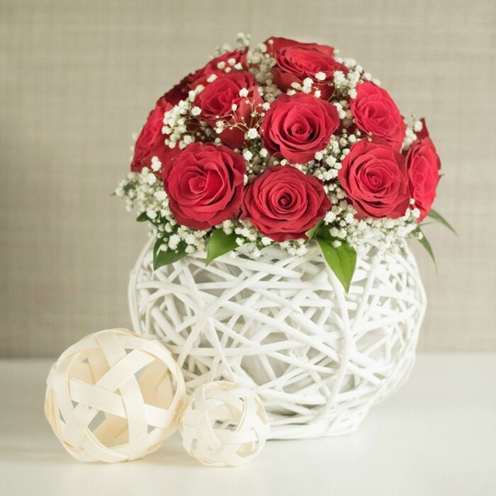 A beautifully styled floral arrangement is presented as if it were the centrepiece of a wedding table set somewhere near Wimbledon Chase or along the leafy streets of Merton. The composition is anchored by a lush, rounded bouquet of deep red roses, each flower open at its fullest, with layered petals that form a velvety spiral from the centre to the outer edges. The roses are positioned closely together, creating a rich, continuous dome of crimson. Nestled between them are clusters of white baby's breath, their tiny, cloud-like blooms offering a delicate contrast and adding a sense of air and lightness to the arrangement. A few discreet green leaves appear around the base and between stems, giving a natural, garden-grown feel that suggests the flowers were freshly arranged at a local florist's workbench. This bouquet rises from a distinctive, round white vase, its surface formed entirely from a network of interwoven strands that cross and loop to create a bulbous, open-lattice structure. The woven design feels both modern and handcrafted, the kind of piece you might find decorating an intimate reception venue near Morden or a stylish home overlooking Cannon Hill Common. To the left of the vase, resting on the same pale surface, are two decorative spheres in soft cream tones. They are also made from woven strips, their open, airy construction echoing the vase's pattern; one is larger and placed slightly forward, while the smaller sits behind, adding gentle depth and symmetry. Behind the scene stretches a softly textured wall in neutral beige, subtly blurred so the vivid red roses and crisp white elements remain the focal point. Soft, even lighting bathes the composition, creating an atmosphere of calm celebration and understated elegance, ideal for weddings, anniversaries, or special dinners in and around Merton.