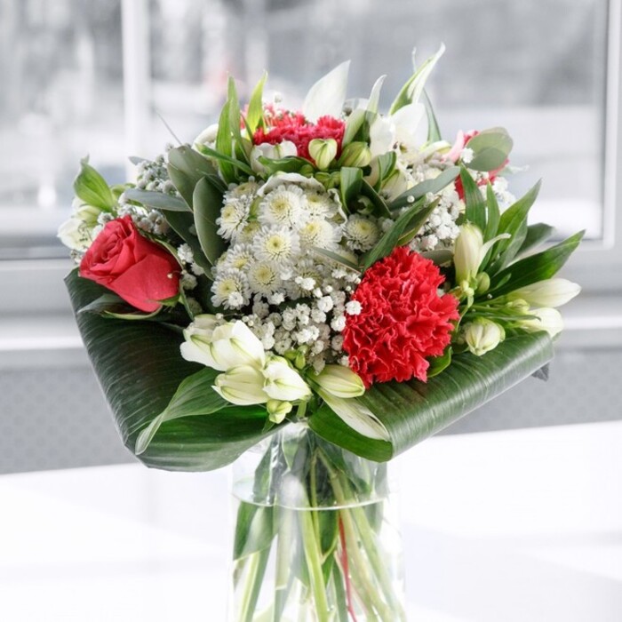 A lush, contemporary bouquet of red and white flowers commands attention from the centre of a sleek cylindrical glass vase, its stems perfectly aligned in clear water as though freshly arranged in a Merton kitchen flooded with daylight. The primary impression is of rich, romantic colour: deep red roses sit proudly at different heights, their velvety petals spiralling outward with that classic, layered swirl that speaks of love and devotion. At the front, a single, densely ruffled crimson carnation provides a focal point, its textured petals folding in on themselves like soft fabric, adding drama and depth to the arrangement. Supporting these bold statements are a series of white blooms that introduce calm and balance-elegant white alstroemeria with slender petals and faint inner markings, soft creamy flowers akin to chrysanthemums or daisies, and several graceful white lily buds still tightly closed, hinting at future blossoms and enduring beauty. Fine sprays of white gypsophila, or baby's breath, drift between the larger flowers like a light mist, giving the bouquet an airy, romantic feeling often chosen for anniversaries or heartfelt congratulations around Wimbledon, South Merton, and nearby streets. The greenery is just as carefully considered: broad, glossy dark green leaves are wrapped and curled neatly around the base, forming a firm, sculptural collar that holds the composition together. Above, a mixture of eucalyptus-like sprigs and slim, pointed leaves adds a play of shapes and green tones, from deep forest hues to softer, silvery greens. Resting on a spotless white surface and framed by a softly blurred, bright background-perhaps a pale wall or a large window-the bouquet glows in even, natural light. It feels both polished and welcoming, the sort of floral centrepiece a local florist might recommend for a special dinner, a thoughtful thank-you, or simply to bring a touch of refined warmth to a Merton home or office.
