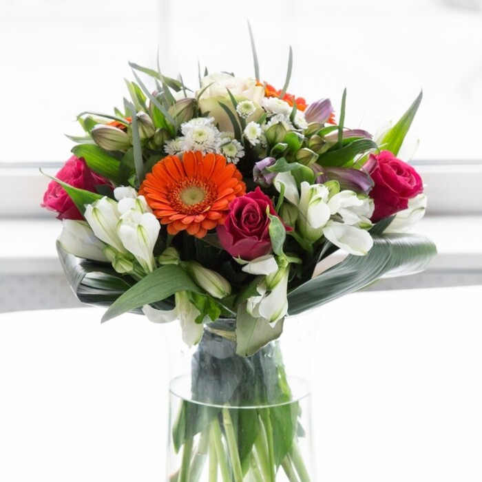 In this exuberant arrangement, the unmistakable freshness and artistry of Merton's finest floristry comes alive. Four prominent gerberas-two in bold orange and two in a gentle peach-offer a radiant focal point, their daisy-like petals curling outwards, surrounding chocolate-brown centers that evoke the warmth of a community gathering in John Innes Park. Among them nestle sumptuous magenta roses, their spirals hinting at layers of affection and grace. Clusters of white alstroemeria and petite spray roses weave lightness and brightness through the bouquet, fragrant and uplifting, while occasional soft pink florets add a tender undertone. Wrapping the whole display, lush aspidistra leaves arc protectively around the base, grounding the arrangement with a touch of garden-inspired elegance-mirroring the abundant greenery seen around Merton's neighbourhoods. Finer sprigs and pointed leaves tuck between the blooms, delivering a sense of fullness and harmony. The clear glass vase stands tall and discreet, showcasing verdant stems and anchoring the composition's organic appeal. Bathed in soft, ambient light, each blossom radiates vitality-a loving choice for new beginnings, celebrations, or simply bringing the serene essence of Merton's local charm into one's home.