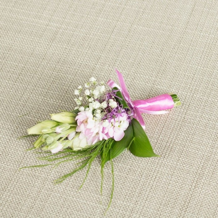 A meticulously crafted floral boutonniere or petite corsage is presented in the centre of the frame, resting on a finely woven neutral linen in a soft greige tone, similar to the table linens you might see at a wedding reception in a Merton venue near Wimbledon Common. The fabric's faint cross-hatched texture provides a calm, organic backdrop that gently contrasts with the vivid greens and romantic pastels of the flowers. At the heart of the arrangement, several soft pink, multi-petalled blooms-resembling delicate stock or small ruffled garden flowers-cluster together in a rounded form. Their petals show subtle gradations of colour, from muted rose at the inner whorls to paler, almost powder-pink edges, giving the cluster a softly luminous quality. Woven between these central blossoms are numerous tiny, snow-white baby's breath flowers, their minute, cloud-like clusters adding an airy, almost ethereal texture and a hint of traditional bridal charm. Tucked here and there among the pink and white are slender spikes of lavender or light purple florets, providing a thoughtful hint of contrast that deepens the palette without disturbing its gentleness. Extending gracefully from one side of the design are several closed buds in creamy white touched with pale green, similar to freesias or other slim, tubular blooms just waiting to open, evoking a sense of anticipation, like the minutes before a ceremony begins. The whole cluster is anchored by glossy, broad green leaves that form a bold, emerald base, while finer, grass-like fronds and ferny sprigs arc outward in delicate wisps, lending a natural, garden-inspired feel. At the base, the stems are neatly aligned and tightly wrapped in a vivid fuchsia satin ribbon, tied into an elegant bow with gently looping ends that rest softly on the linen. The light falls evenly across the composition, enhancing the freshness of each petal and leaf and capturing a floral accessory that would sit beautifully on a lapel or wrist at any Merton wedding, prom, or special celebration, subtly reflecting both craftsmanship and local style.