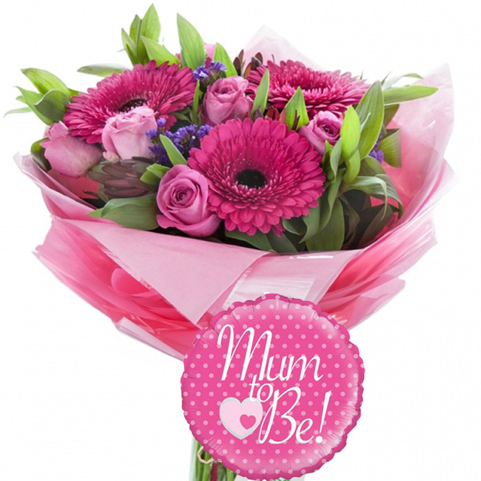 This Mom to Be floral gift is presented against a bright white background, allowing every colour and texture to stand out clearly, much like a fresh bouquet laid out for inspection in a Merton Flowers studio. Dominating the arrangement are several large fuchsia gerbera daisies, each flower a flat, circular burst of colour with a dark central eye and layers of neat, radiating petals that suggest happiness and energy. Threaded delicately between these bold blooms are pastel pink roses, offering a contrast in both shape and mood. Some of the roses are closed into rounded buds, promising future beauty, while others are beginning to open, revealing concentric rings of soft, velvety petals that curl gently at their edges. Small clusters of purple filler flowers, possibly statice or limonium, nestle in the gaps, creating a soft haze of cooler colour and adding a slightly airy texture. The overall shape of the bouquet is rounded and full, with different heights and angles giving it a natural, hand-tied feel, as if it has just been finished for same-day delivery across Merton and neighbouring Wimbledon. Around the flowers, a variety of fresh green foliage adds depth and structure: long, slender leaves arc gently outwards, while broader glossy leaves peek from within the bouquet, providing a rich green frame for the brighter petals. The stems are wrapped together with multiple layers of pink tissue paper in both blush and stronger tones, forming a voluminous, ruffled collar at the base. A final layer of clear, shiny cellophane encases the tissue, catching and reflecting light in a way that enhances the sense of celebration. Rising above the blooms is a round fuchsia foil balloon edged with subtle scalloping and decorated with crisp white polka dots. The balloon bears the joyful message Mom to Be! in graceful white script, complete with a sweet double-heart motif, making this image a perfect visual for baby showers, pregnancy reveals, or heartfelt congratulations for expectant mothers across the Merton area.