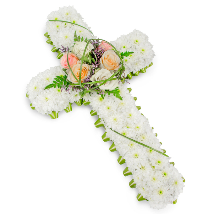 A traditional Christian floral cross is shown in elegant detail, resting against a clean white background that enhances its calm, reverent character-much like a tribute you might encounter in a Merton church or at a quiet crematorium service near Mitcham. The structure of the cross is strong and clearly defined, yet its surface is completely softened by a dense overlay of small, immaculate white chrysanthemums or similar blooms. Each flower head is carefully nestled into place so that, viewed as a whole, the cross resembles a plush, gently textured cushion of white petals. This continuous floral covering conveys purity, consolation, and a sense of serene farewell. Running along the perimeter is a slim but striking border of lush green foliage, perhaps folded leaves or a specialist floral edge, that outlines the cross with precision. The bright green trim adds contrast and frames the white flowers, echoing the natural greenery of local parks and commons around Merton. At the centre, where the cross's arms meet, a prominent, slightly elevated spray of roses forms the emotional heart of the design. Here, larger blooms open in soothing shades of soft peach, apricot, delicate blush pink, and creamy ivory, each rose layered and full, suggesting warmth, gratitude, and remembrance. These focal flowers are surrounded by airy fern fronds, slender, arched strands of lime-green grass, and discreet flecks of purple filler flowers that add both texture and a gentle hint of colour. The overall lighting is soft but clear, allowing every petal and leaf to be appreciated. This floral cross stands as a classic funeral tribute for families across Merton, offering a visual expression of faith, love, and enduring connection.