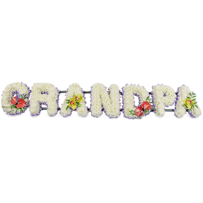 Against a clean, uncluttered white background, a long funeral tribute spells out GRANDPA in bold block capitals, stretching from left to right in a continuous arrangement. Each letter is transformed into a padded floral form, completely covered in fresh white chrysanthemums. The small, uniform blooms are packed into every space, creating a smooth yet slightly textured surface that looks plush and full, like a soft cushion of petals. Their bright white hue suggests purity and remembrance, making the tribute feel both dignified and gentle. Along the base of each letter, a narrow band of pale lilac ribbon can be seen, adding a delicate line of colour that outlines the shapes without disturbing the simplicity of the design. Hidden dark green bars support the letters from behind, giving the tribute the strength to sit securely on an easel or stand during a funeral in the Merton area, whether at a church near South Wimbledon or a crematorium chapel. Carefully placed accent sprays punctuate the word. On the G and the central D, clusters of rich red anthuriums are arranged among deep green foliage. Their glossy, heart-shaped petals and upright yellow centres draw the eye and suggest deep love and gratitude. On the first A and the final A, lime-toned orchids are arranged in graceful arcs, their elegant, star-like blooms and verdant leaves bringing a contemporary, uplifting feel, as if acknowledging both sorrow and cherished memories. The overall impression is of a bespoke memorial arrangement prepared by an experienced local florist, offering a gentle yet clearly spoken tribute to a much-loved grandfather from Merton.