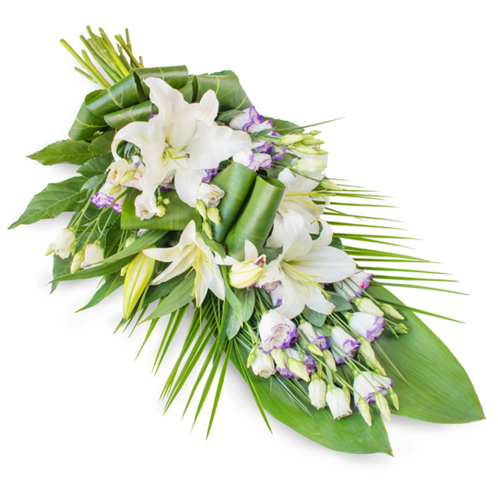 A long, teardrop-style funeral spray lies diagonally across a stark white background, composed in soothing tones of white, green, and a whisper of lavender, as if prepared for a quiet farewell at a Merton chapel or cemetery. Dominating the arrangement are white oriental lilies, their large, trumpet-shaped flowers opening in stages. Some lilies are fully unfurled, with thick, velvety petals arching back to frame gentle yellow-green stamens; others remain partially open or still in elegant, pointed buds, suggesting both memory and hope. Interspersed between these lilies, clusters of lisianthus flowers add delicacy and movement. Their petals are soft white, with edges tinged and veined in light purple, bringing a subtle contrast that never disturbs the calm mood of the piece. Small, unopened lisianthus buds and stems weave throughout the spray, creating fine points of interest for the eye to follow. Supporting this floral canopy is a generous foundation of lush foliage. Broad, dark green leaves form a glossy backdrop, some left flat to anchor the design, others meticulously folded, curled, and rolled into sculptural loops that demonstrate experienced handiwork, like something crafted in a long-established Merton flower shop. From one side and the lower portion, long, slender palm-like fronds extend outward, echoing the overall diagonal line and helping the spray taper to a gentle point. The greens vary from deep forest tones to bright, fresh hues, symbolising life and renewal around the serene white blooms. The arrangement conveys a sense of dignified peace and heartfelt remembrance, ideal for casket adornment, graveside tributes, or memorial displays across Merton, Wimbledon, and nearby neighbourhoods.