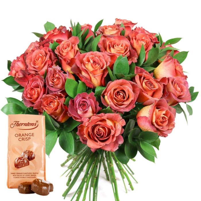 Dominating the center of the image is a sumptuous bouquet of fresh roses, expertly arranged by local hands and backed by the crisp clarity of a white background. The roses delight in a spectrum of hues: from delicate peach and golden orange at their cores, to lively coral, deepening rose-pink, and near-red at the outer petals. This ombré coloration, coupled with densely layered, velvety petals, imparts volume and opulence akin to floral displays in Merton's village markets. Between the abundant blooms, dark, verdant foliage-perhaps salal or ruscus, both favoured by florists in Merton-provides a structured, natural frame, enhancing the vibrancy and depth of the overall design. In the lower left, supporting this floral showpiece, a Thorntons Orange Crisp chocolate pouch in pale cream stands upright, elegantly printed with the iconic logo and product description, illustrated by two smooth, dark chocolates. In front, a pair of unwrapped glossy truffles gleam, their deep caramel sheen a testament to their richness. The bouquet's stems are neatly gathered, a detail that hints at the bouquet's hand-tied craftsmanship and readiness for prompt delivery throughout Merton and Wimbledon. The entirety is illuminated by soft, natural light, ensuring every shade and texture is captured, making the scene ideal for commemorating birthdays, anniversaries, Valentine's Day, or moments of appreciation with a distinct Merton flair.