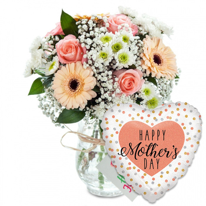 Set against a bright white background, this Mother's Day ensemble looks composed and thoughtful, as if waiting on a tidy hall table in a Merton home. On the left sits a fresh floral bouquet arranged in a clear, slightly jar-shaped glass vase, its rounded form mirroring the generous dome of flowers above. The narrow neck of the vase is wrapped with a simple piece of raffia-like twine tied into a bow, lending a relaxed, handmade quality that suits the friendly, residential streets of Raynes Park and South Wimbledon. The bouquet itself is full and lush: several soft peach and blush-pink roses are shown at varying stages of bloom, from gently unfurling petals to fully open spiral rosettes. Alongside them, two large peach gerbera daisies stand out with their dark brown centres framed by numerous smooth, radiating petals, adding a bolder, contemporary note. Scattered throughout the arrangement, clusters of fine white baby's breath bring a delicate, cloud-like texture, while smaller white chrysanthemum heads with pale green centres fill the gaps, making the bouquet look rounded and complete. Shiny green leaves arch between the blooms, deepening the colour palette and contributing a fresh, garden-picked character. To the lower right, overlapping the front of the vase, rests a festive heart-shaped foil balloon. Its white surface is speckled with playful gold and silver dots, like confetti from a family celebration in a Merton park. At the centre of the balloon, a peach or light coral heart holds the message HAPPY Mother's DAY printed in black, with HAPPY in a modern block font and Mother's DAY in an elegant cursive style. A small corner of a white greeting card peeks between the flowers and balloon, hinting at a personal note. The overall image suggests a complete Mother's Day surprise, perfect for doorstep delivery across Merton, Wimbledon, and the surrounding areas.