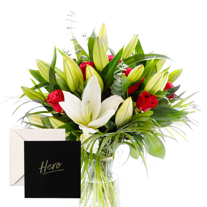 As a florist, I love blending bold with serene, and this arrangement does so with effortless grace. The white lilies drift toward the heart of the bouquet, luminous and airy, their satin petals surrounded by lush, forest-green foliage. Rich ruby roses punctuate the design, each bloom a symbol of love and respect. The verdant leaves provide a cooling backdrop, while unopened lily buds hint at new beginnings on the journey ahead. In soft, natural light the bouquet feels alive, its textures and tones catching every nuance of shade and shine. It is an honouring gesture, ideal for marking a milestone, a celebration, or a heartfelt thank you. The accompanying card reads Hero, lending intimacy and staying power to this enduring tribute. The bouquet rests in a clear glass vase, water visible through the glass, stems arranged with careful spacing to maintain balance. Available for delivery across South Wimbledon, Colliers Wood, and surrounding Merton communities, this creation offers artistry, warmth, and a sense of lasting gratitude. It is a gift that speaks of strength and achievement, wrapped in beauty and poised for impact.