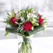 A striking red and white bouquet stands proudly in a clear cylindrical glass vase, its stems neatly trimmed and fanned out in fresh water, like something you might see in a sunlit front room off Wimbledon Chase in Merton. The arrangement is dominated by velvety deep red roses, each bloom perfectly formed with petals just beginning to unfurl, echoing a timeless sense of romance and heartfelt passion. At the very front, a large crimson carnation, densely ruffled and layered, becomes a rich textural focal point, drawing the eye into the heart of the display. Around it, smaller red blooms and accents carry the colour through the design, creating a steady rhythm of scarlet tones. Interlaced among the reds are crisp white alstroemeria, their slim petals delicately streaked and speckled at the centre, along with creamy white blooms that resemble daisies or chrysanthemums. Several white lily buds remain tightly closed, their long, tapered forms suggesting promise and future beauty, much like a special celebration still unfolding. Tiny clusters of white gypsophila, or baby's breath, float like soft clouds among the bolder flowers, adding airy lightness and a feeling of generosity. The bouquet is cradled by lush greenery: glossy dark green leaves are artfully curled and wrapped around the base, forming an elegant collar, while eucalyptus-like sprigs and pointed foliage add depth and a variety of green tones. The vase sits on a pristine white surface beneath even, natural light, as though placed near a bright window in a Merton townhouse, the background softly blurred to keep focus on the flowers. Overall, the mood is one of thoughtful celebration-ideal for anniversaries, birthdays, Mother's Day, or a warm, elegant gift to brighten a home or office anywhere across the Merton area.