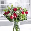 A lush bouquet of deep crimson red roses and matching ruffled carnations stands proudly in a clear glass vase on a bright white tabletop, bathed in gentle daylight from a large, softly blurred window behind. The roses, some fully open with velvety petals spiralling outward and others still in bud, are clustered at the heart of the arrangement, their rich colour evoking romance and heartfelt celebration. Nestled between them are pristine white alstroemeria, many still in tight, promising buds, with a few trumpet-shaped blooms just beginning to open, their petals tipped with the faintest hint of green. Fine sprays of tiny white gypsophila, or baby's breath, thread through the bouquet like a soft mist, adding an airy lightness that contrasts the bold red blooms. Around the base and rising through the centre, broad, glossy green leaves form a natural collar, while slender, more delicate greenery winds upward, lending movement and texture. The stems are neatly cut and clearly visible through the water in the vase, reinforcing the sense of freshness, as if the flowers were just arranged by a careful florist in Merton that morning. The white tabletop reflects a subtle glow back onto the vase, while the muted, almost monochrome view beyond the window keeps all attention fixed on the vivid reds, clean whites, and rich greens. The overall mood is one of joyful elegance, ideal for a birthday, anniversary, or simply to brighten a home off London Road or near Morden Hall Park with a touch of classic floral beauty.